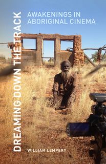 Bearded man sits among dried grasses before a roofless ruin, the scene captured by a movie camera in the foreground. Title vertical on left.