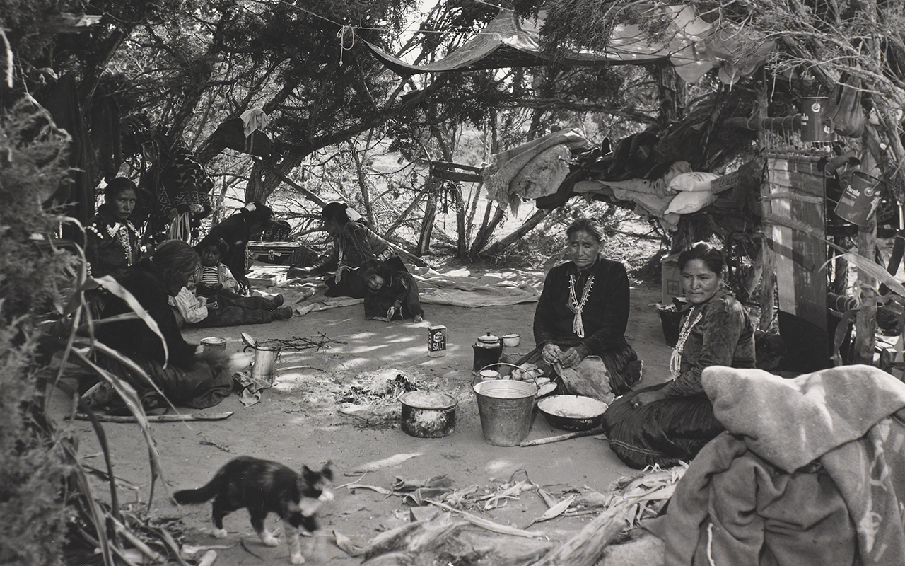 Nine family members, women and children, sit inside a shelter in glade of trees, engaged in domestic activities, with a cat in the foreground.