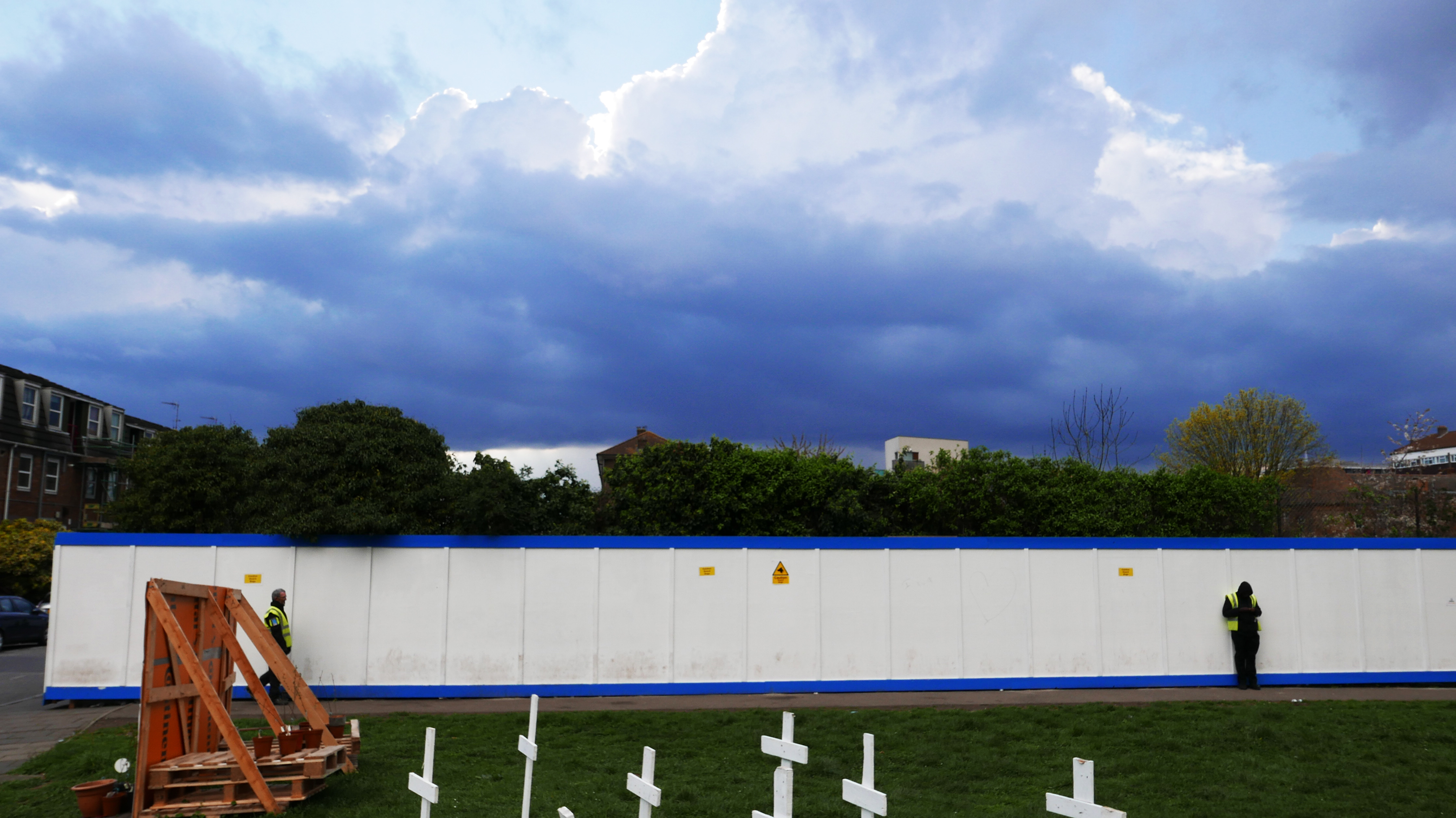 A white wooden wall accented at top and bottom in blue with two security guards stationed before it. White crosses adorn the grassy area before them.
