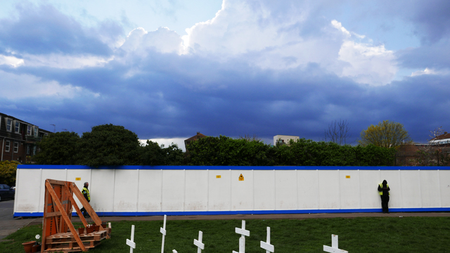 A white wooden wall accented at top and bottom in blue with two security guards stationed before it. White crosses adorn the grassy area before them.