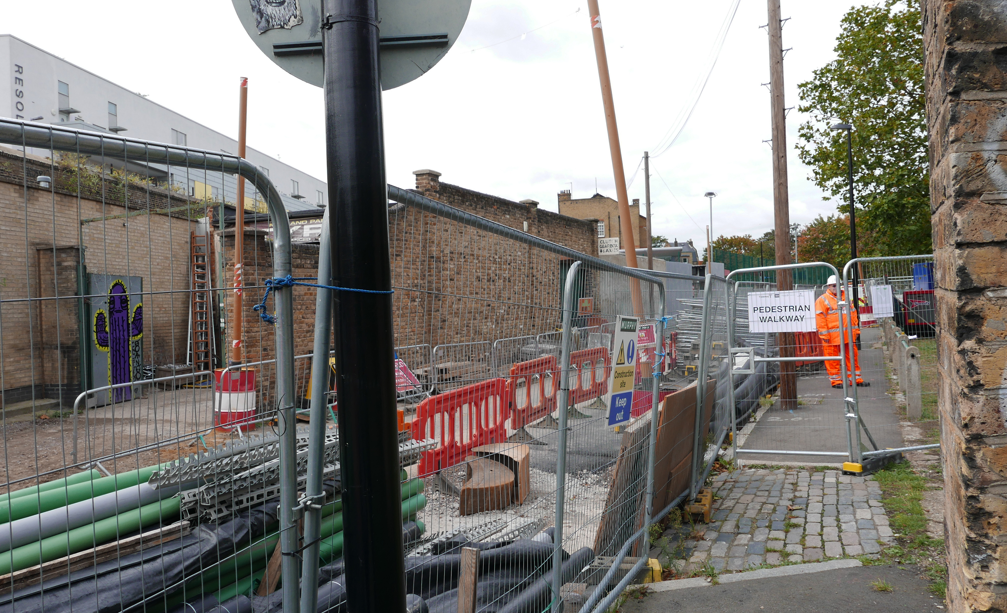 A construction site in an urban setting surrounded by brick buildings. Piping, blockades, and fencing surround the space.