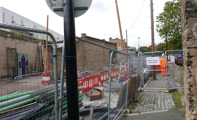 A construction site in an urban setting surrounded by brick buildings. Piping, blockades, and fencing surround the space.