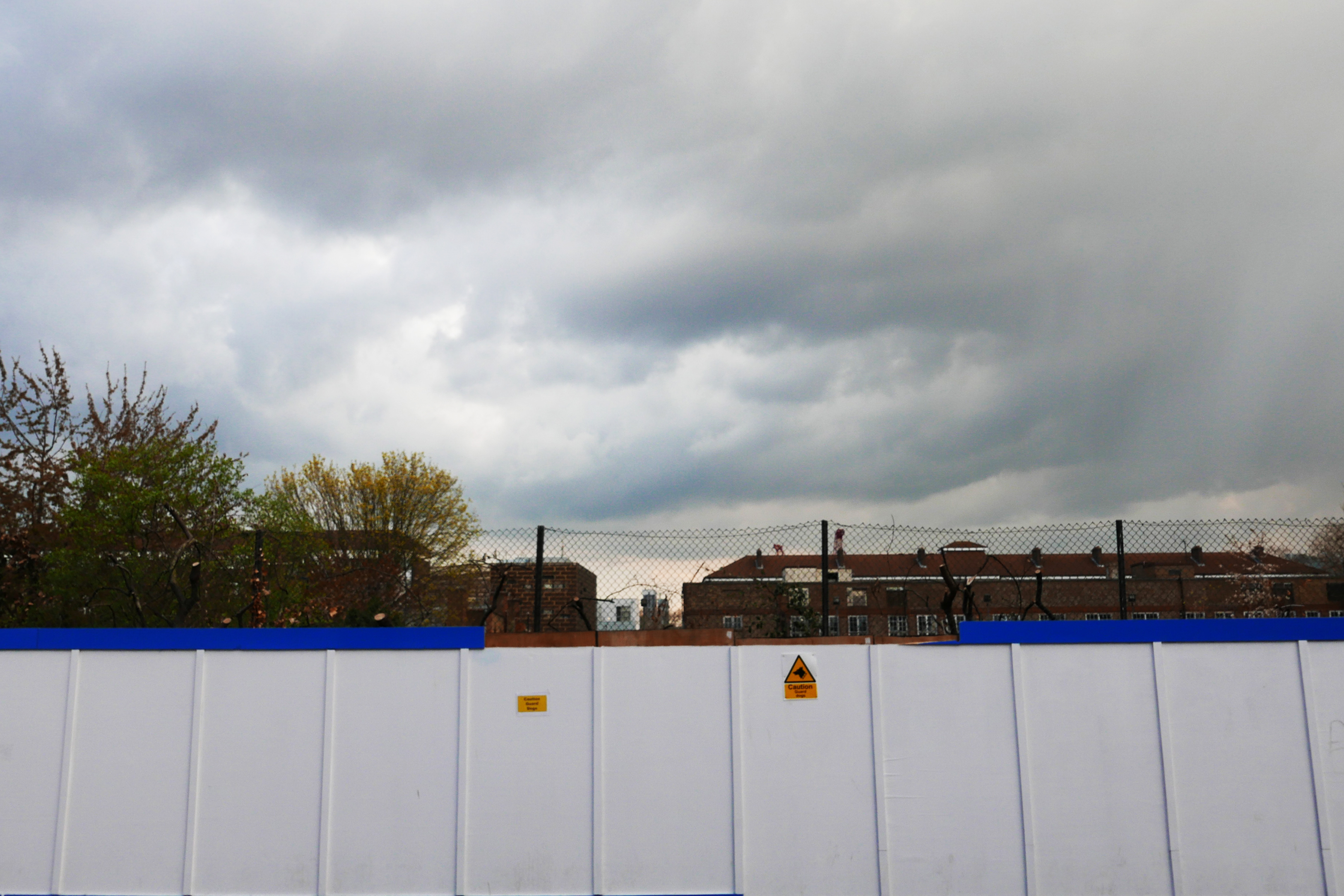A white wooden wall topped with chain-link fencing in the foreground with residential buildings in the background.