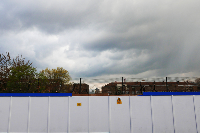 A white wooden wall topped with chain-link fencing in the foreground with residential buildings in the background.