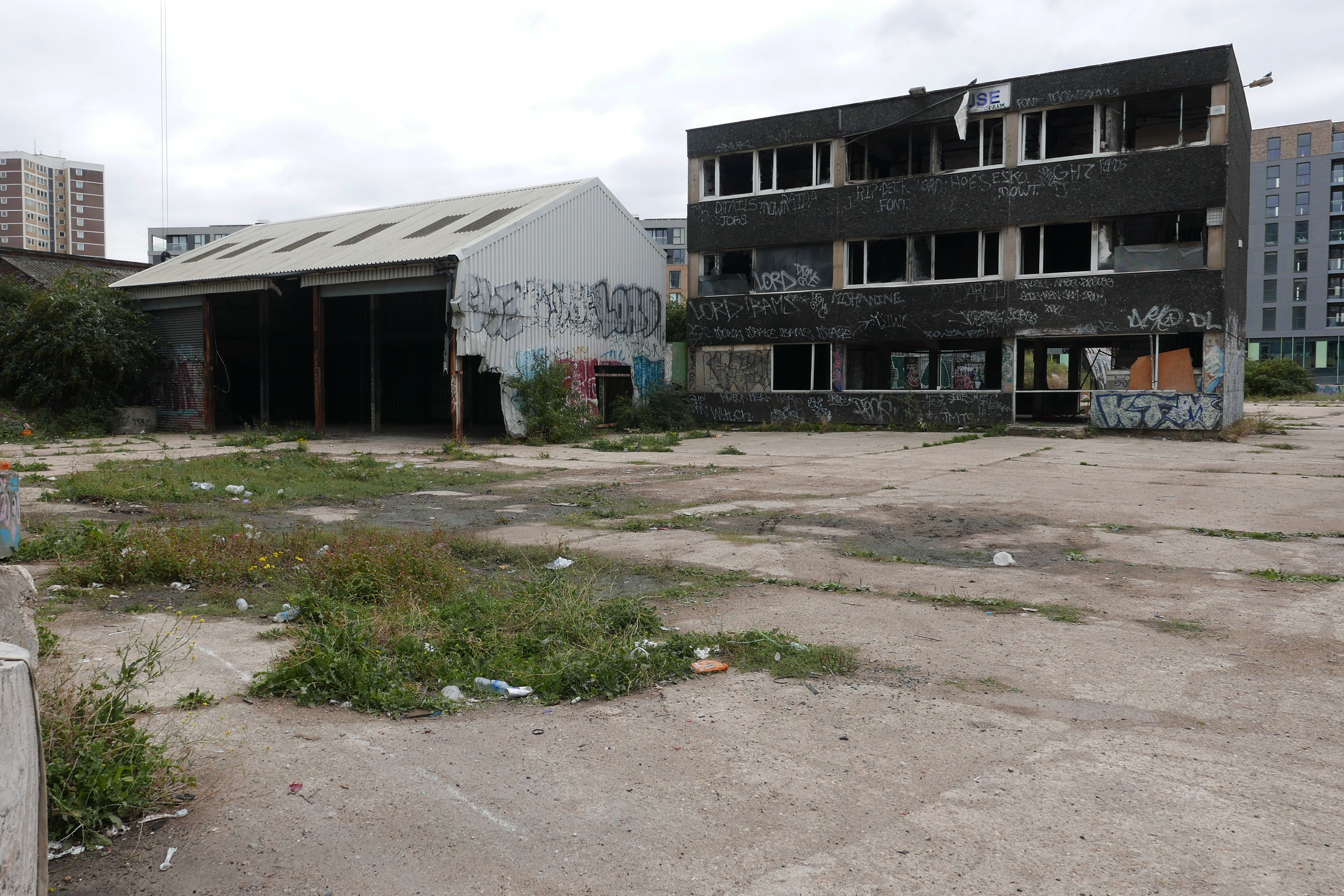 Two small abandoned commercial buildings adorned with graffiti abutting a parking lot littered with weeds and various bottles.