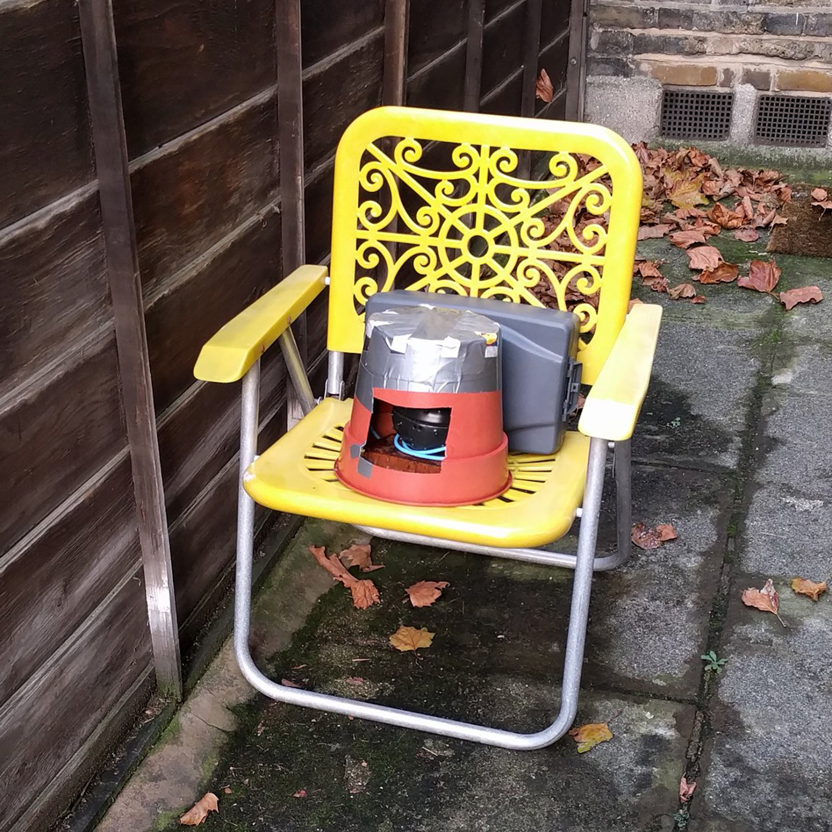 A yellow folding chair upon which a red bucket sits. The bucket has a cutout revealing a sensor array with a black spiny casing.