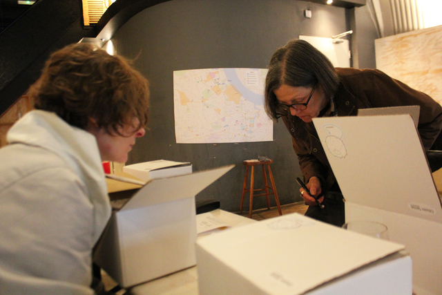 Two people across from one another working at a table covered with white cardboard boxes.