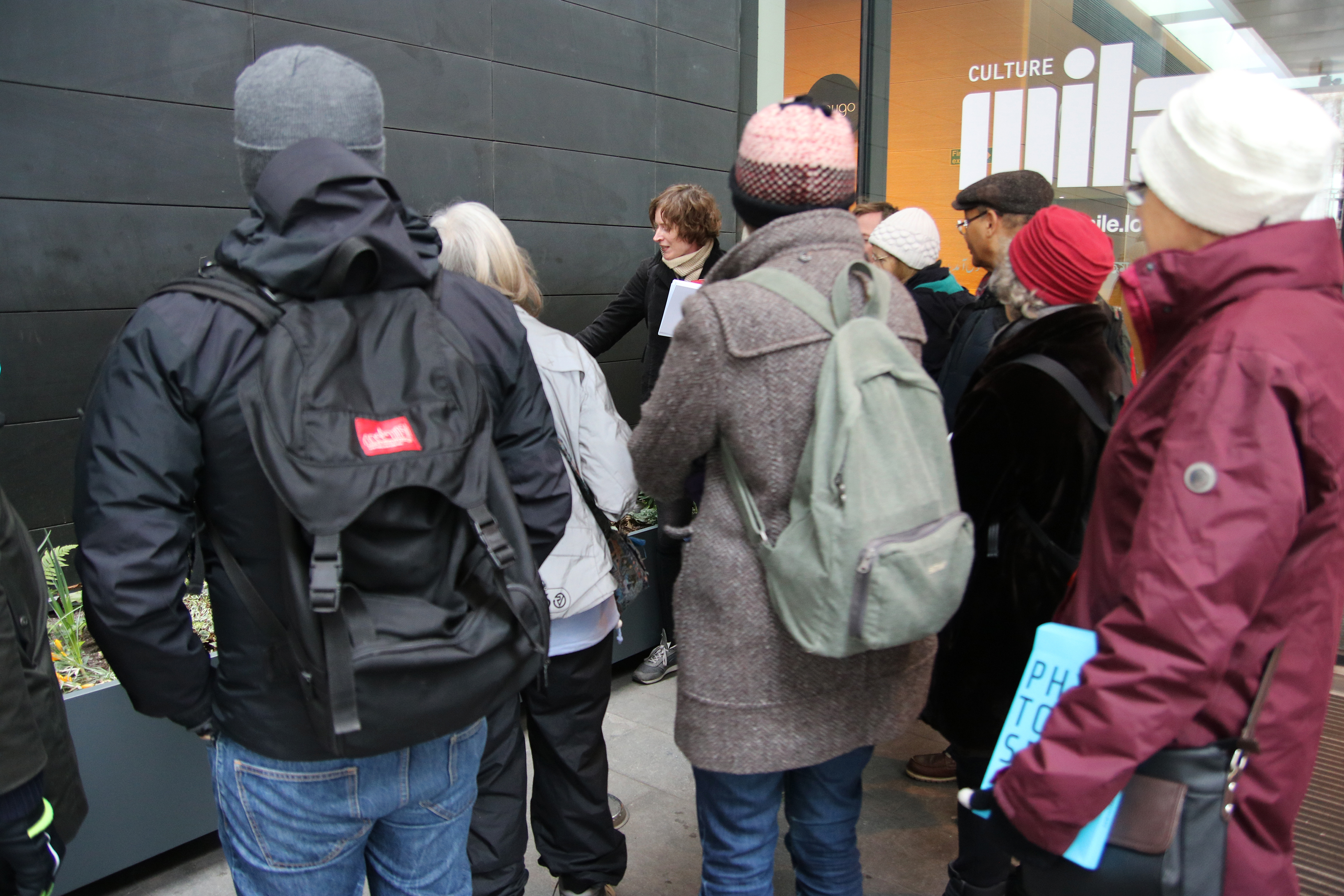A group of people stopped before an above-ground planter, listening to a person speaking beside it.