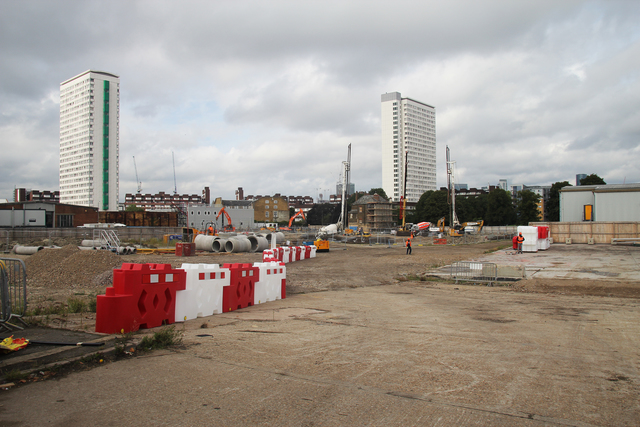 A construction site with red-and-white barricades, concrete cylinders, and various cranes and diggers with personnel scattered throughout.