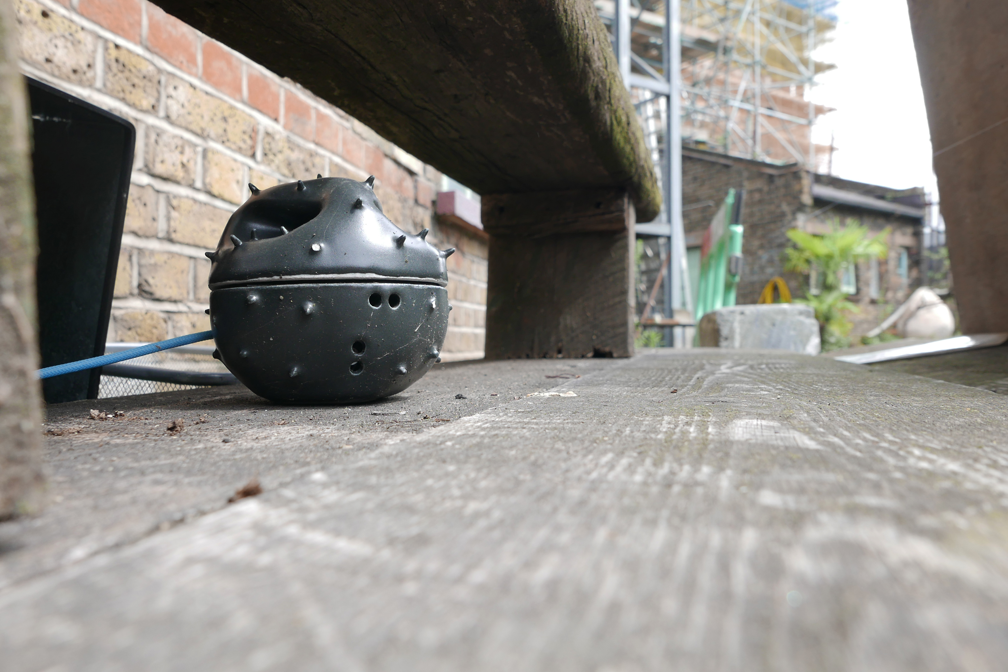 A sensor array enclosed in a black casing with a spiny shell sits on the ground below a wooden bench, trailing a blue cord.