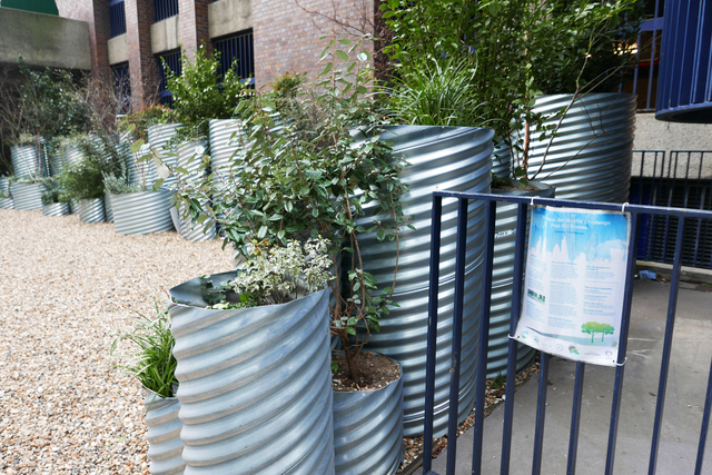 Hollow sheet-metal tubes converted into planters, each holding earth and various plantings within a rocked garden area beside a sidewalk.