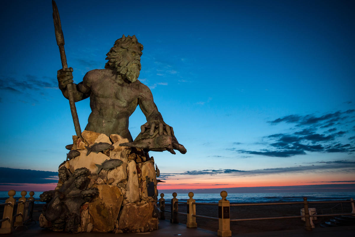 This statue of King Neptune (Poseidon) is depicted having a muscular physique with a beard and long flowing hair, holding a mighty trident in his right hand and holding a sea turtle in his left hand. His body is only shown from the waist up, as the base of the statue is assembled out of large stones. The base is adorned with various depictions of sea life including an octopus, dolphins (other side), small fishes, and starfish.