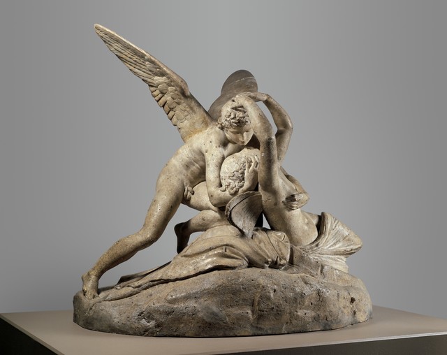 A Neoclassical plaster sculpture of winged god Cupid leaning over a reclining Psyche reaching up around his head in a passionate embrace. The figures are almost kissing with Cupid's wings extended outward and Psyche's head tilted backward. Figures are mounted on a rocky base.