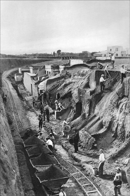A black and white photo of the excavation of Ercolano. A track has been laid down for carts to carry excavated tufo out of the site. People are working the dig.