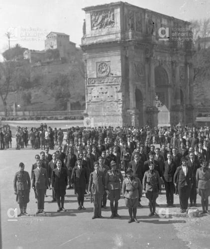 A black and white photo of many uniformed and suited figures standing with intent before the Arch of Constantine in Rome. Among them in Benito Mussolini