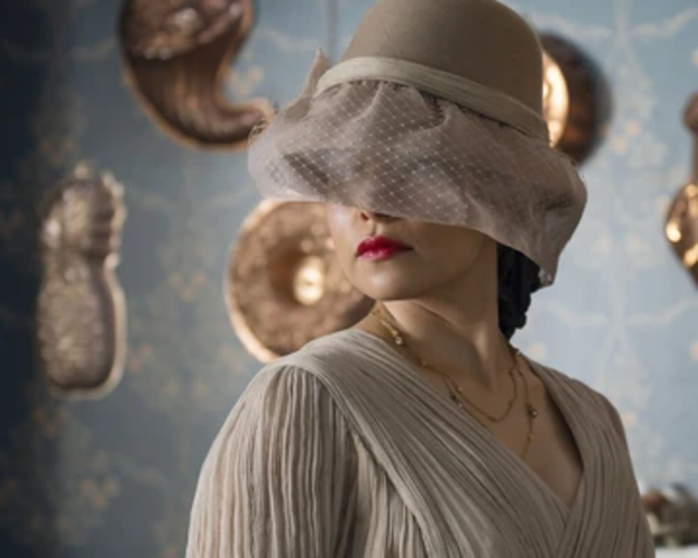 A photo of Medusa, in which she has on a white hat and veil that covers her snake hair and eyes. She is depicted as a beautiful woman, with dark red lipstick.