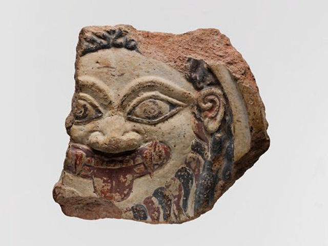 Fragment of an Archaic terracotta antefix depicting the head of Medusa, with large almond-shaped eyes, a broad nose, an open mouth with a protruding tongue, sharp teeth, hair and a beard. Faded red and black paint decorates the facial features and hair. Broken edges show the terracotta clay underneath.
