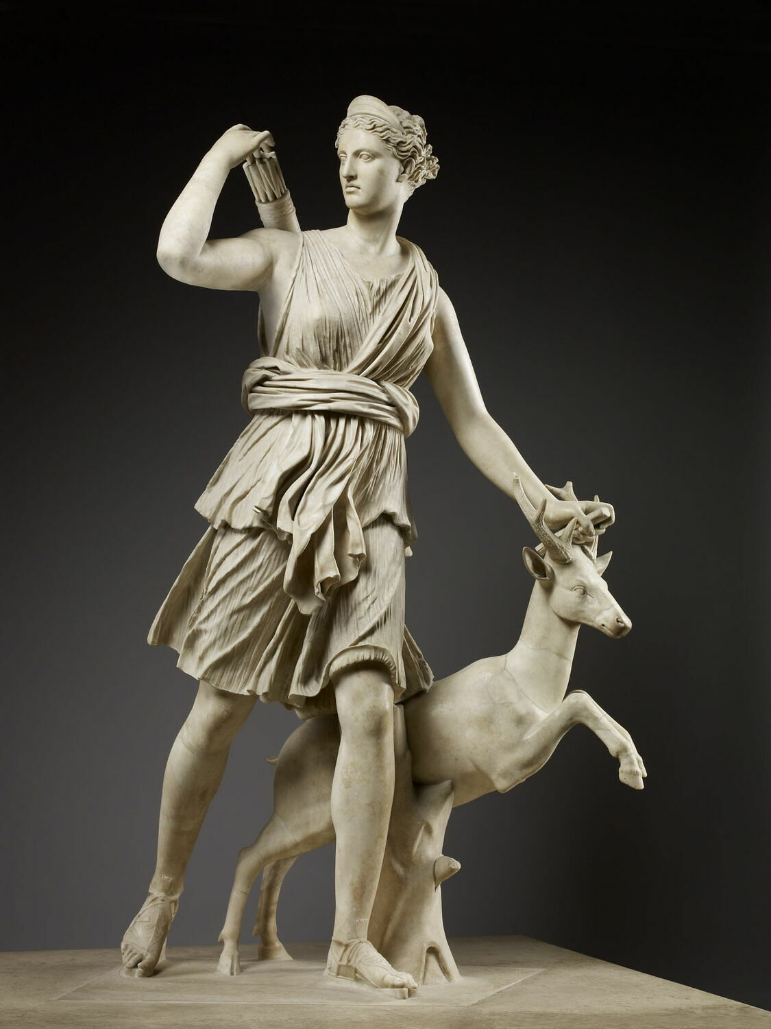 Marble statue picturing goddess Artemis. Poised with her left foot in front, she uses her right hand to reach for a quiver of arrows. Her other hand hovers above the horns of a deer standing on its' hind legs. 