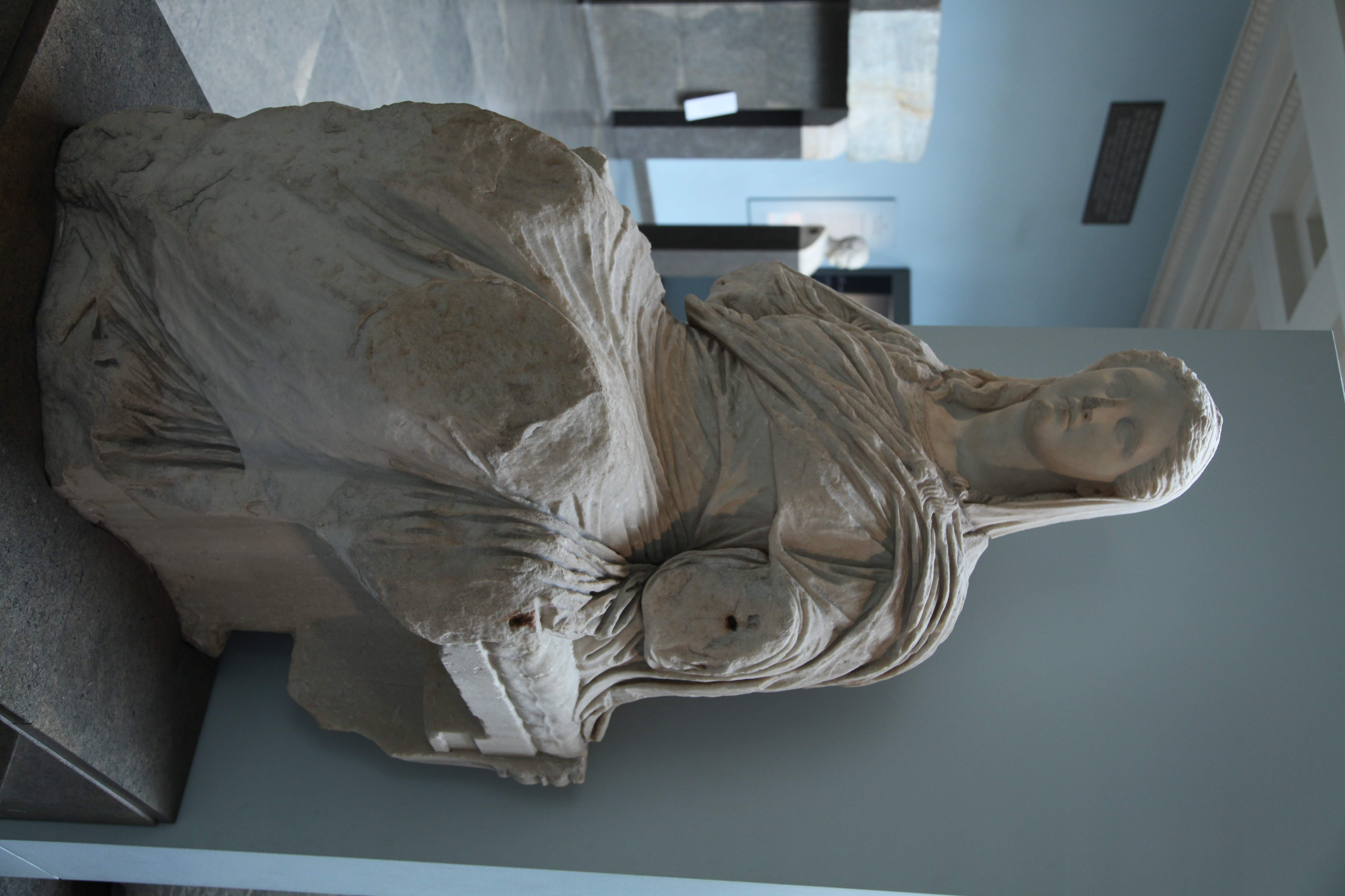 A marble statue of Demeter of Knidos, an ancient Greek sculpture depicting the goddess of agriculture and fertility. She is seated, draped in flowing garments that emphasize the natural folds of the fabric. Her expression is serene, with a gentle gaze and a slightly downturned head. The statue is around 5 feet tall. Though her arms are missing, the craftsmanship suggests a once-majestic presence, in line with the votive offerings the statue would likely receive from worshippers. 