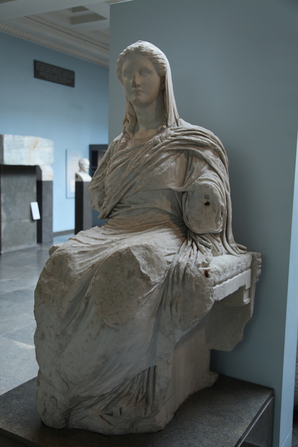 A marble statue of Demeter of Knidos, an ancient Greek sculpture depicting the goddess of agriculture and fertility. She is seated, draped in flowing garments that emphasize the natural folds of the fabric. Her expression is serene, with a gentle gaze and a slightly downturned head. The statue is around 5 feet tall. Though her arms are missing, the craftsmanship suggests a once-majestic presence, in line with the votive offerings the statue would likely receive from worshippers. 