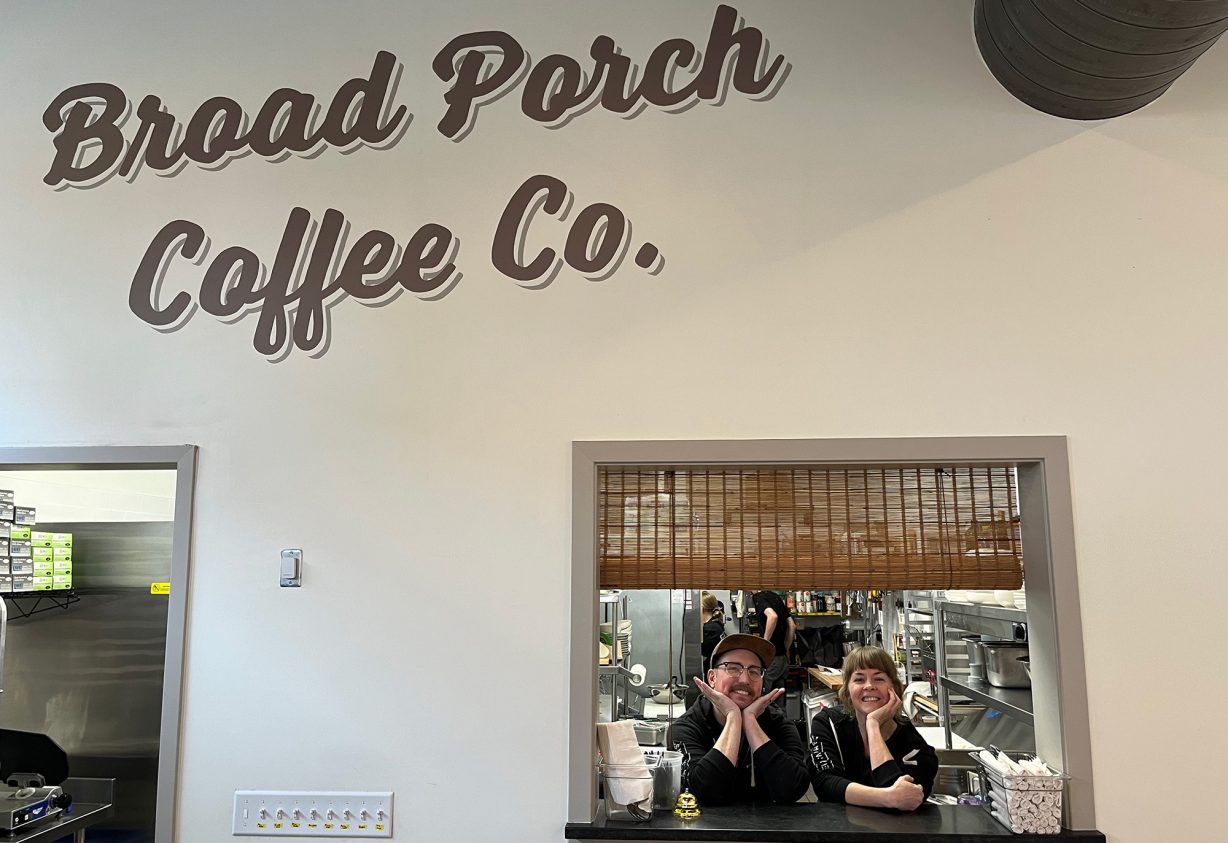 Two people in the opening from a kitchen under a painted sign that says Broad Porch Coffee Co.