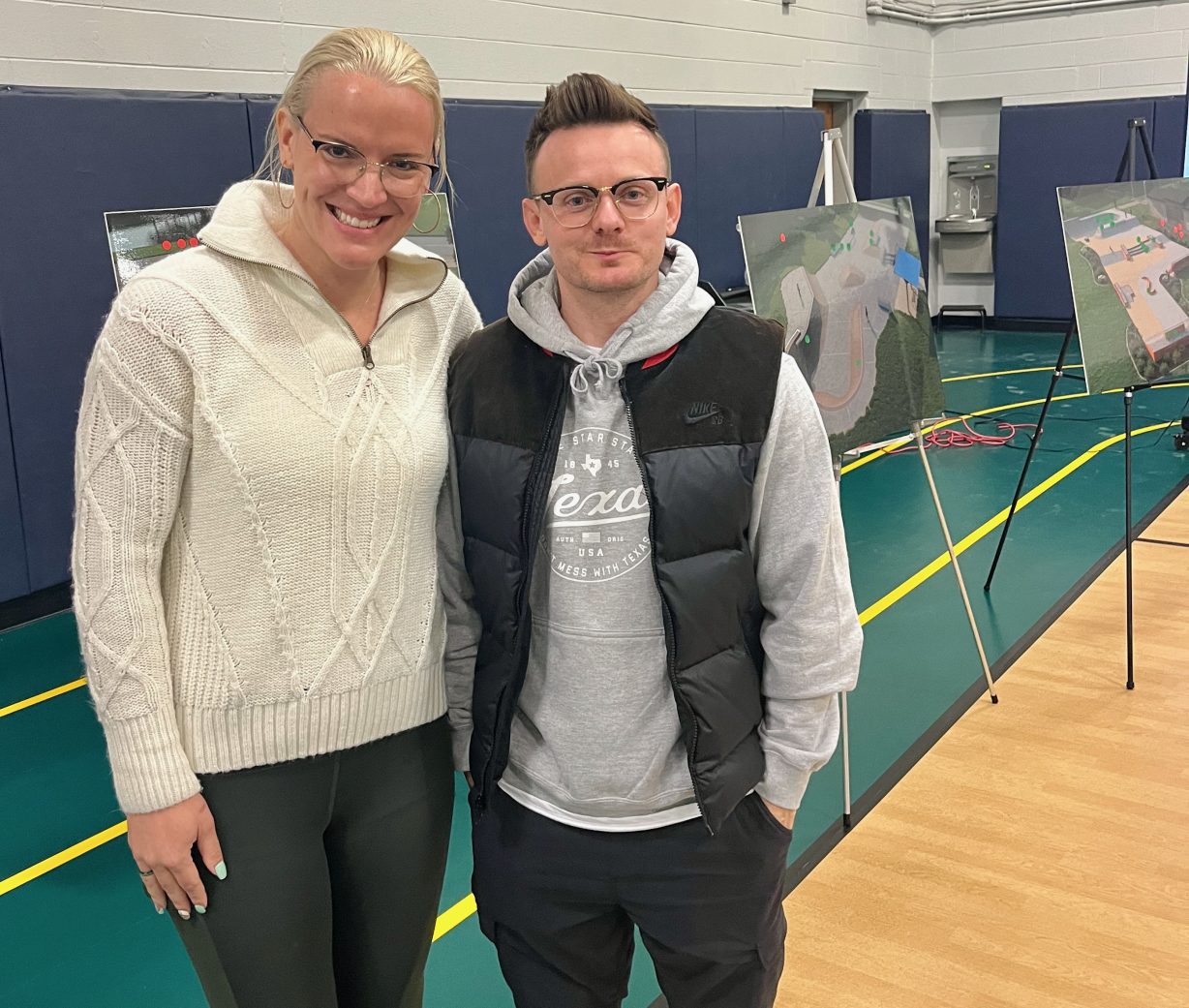 Two people pose for the camera with renderings of skate park designs in the background