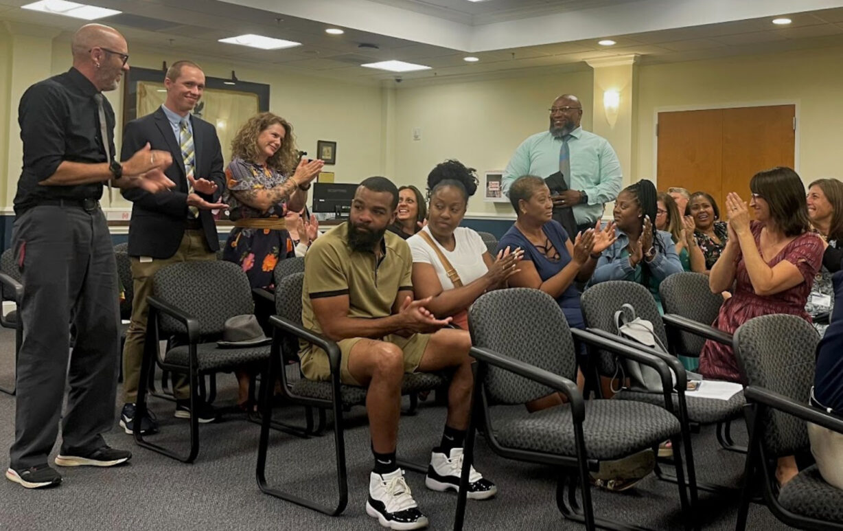 Four people standing while two dozen others sitting clap