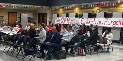 People sitting on folding chairs in a school cafeteria in front of a banner reading "We Support Collective Bargaining"