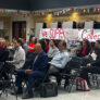 People sitting on folding chairs in a school cafeteria in front of a banner reading "We Support Collective Bargaining"