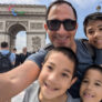 A man taking a selfie with three children in front of the Arc De Triomphe in Paris.