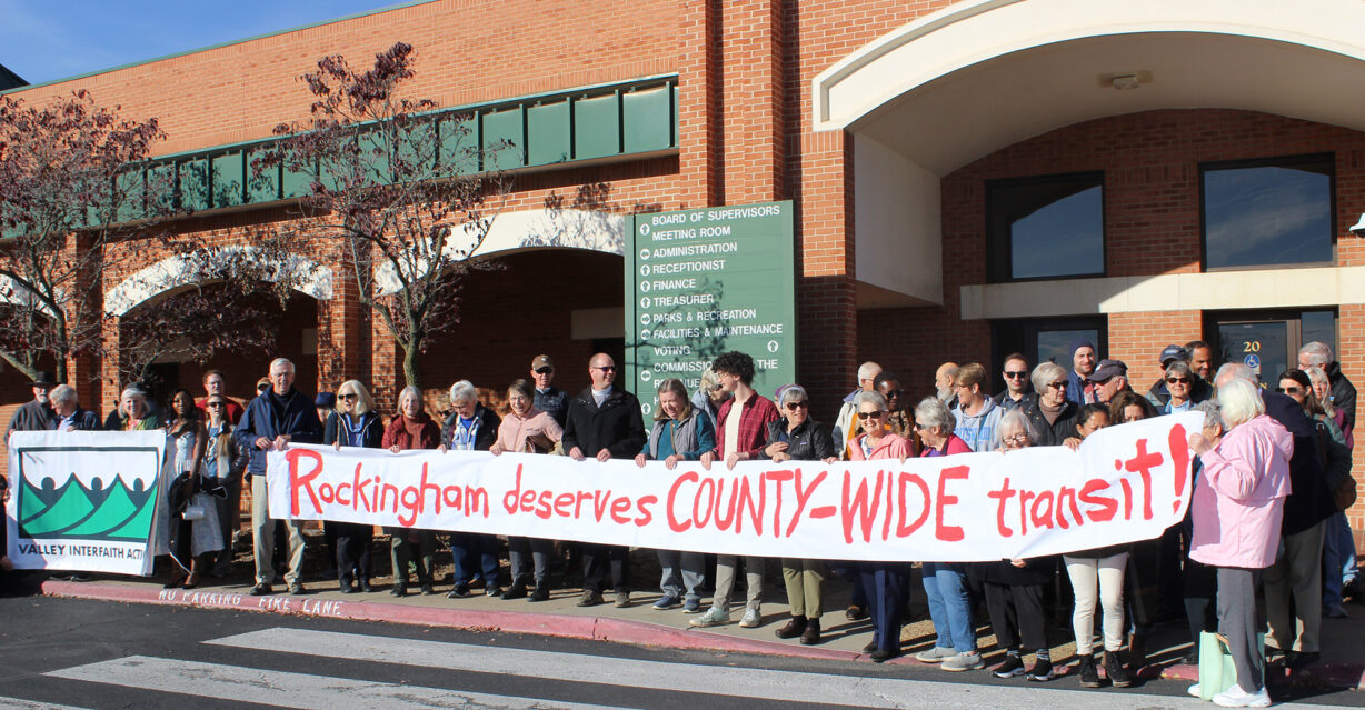 a group of people holding a long homemade sign saying "Rockingham deserves COUNTY-WIDE transit"