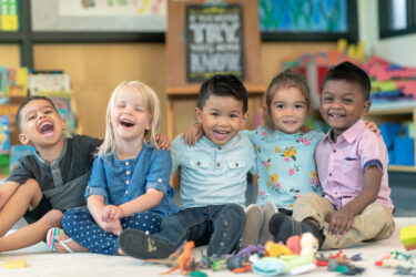 Portrait of a happy multi-ethnic group of preschool students in their classroom. The cute children are sitting in a line with their arms around each other. The kids are laughing and smiling directly at the camera.