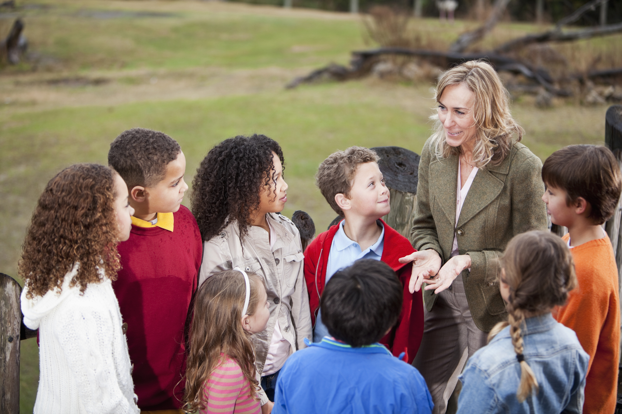 Teacher (50s) with multi-ethnic group of elementary school children at zoo, standing on observation deck overlooking animal exhibit.