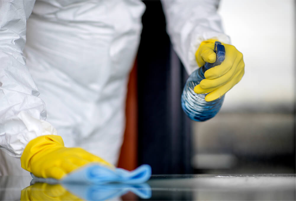 Professional cleaner in white wearing yellow gloves cleaing a surface area with liquid spray bottle and blue rag