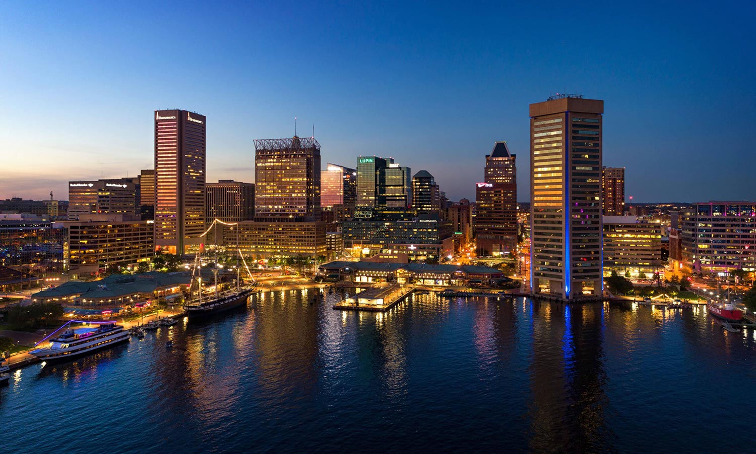 Downtown Baltimore skyline aerial at dusk, with Inner Harbor in the foreground.