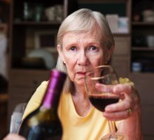 Older adult female offering a bottle of wine and glass sitting alone