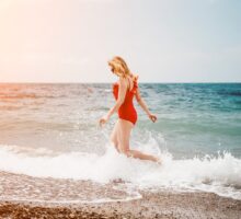 blond woman in red swimsuit walks in water on beach; how to prevent sunburn?