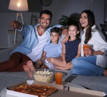 Man, woman and two children sit on sofa with popcorn and pizza on coffee table
