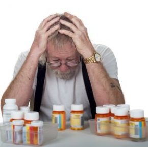 senior sitting behind a lot of pill bottles holding his head in his hands in confusion