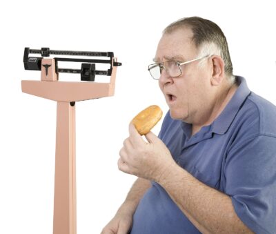 Overweight (obese) older man next to scale eating a donut