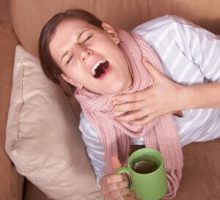 A young woman is sick. She is lying on the couch, sneezing. She has a tea in her hand.