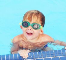 Young boy holding onto side of pool smiling and wearing goggles over his ear