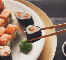 chopsticks holding a piece of sushi with more sushi and a mound of wasabi in the background