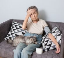 Very old person taking blood pressure with cat on lap and hand at forehead.