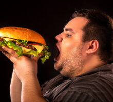 Heavy young man opens mouth wide to bite enormous hamburger