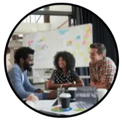 A group of 3 writers collaborate as they sit at a table with a whiteboard of their story structure and character development behind them. 