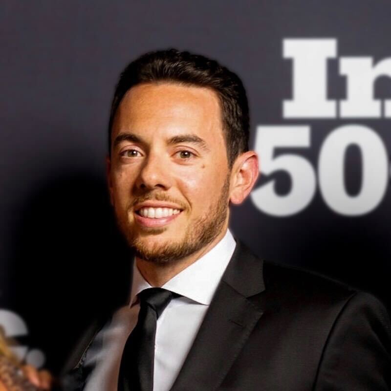 A well-dressed man with short dark hair and a trimmed beard, wearing a black suit and tie.