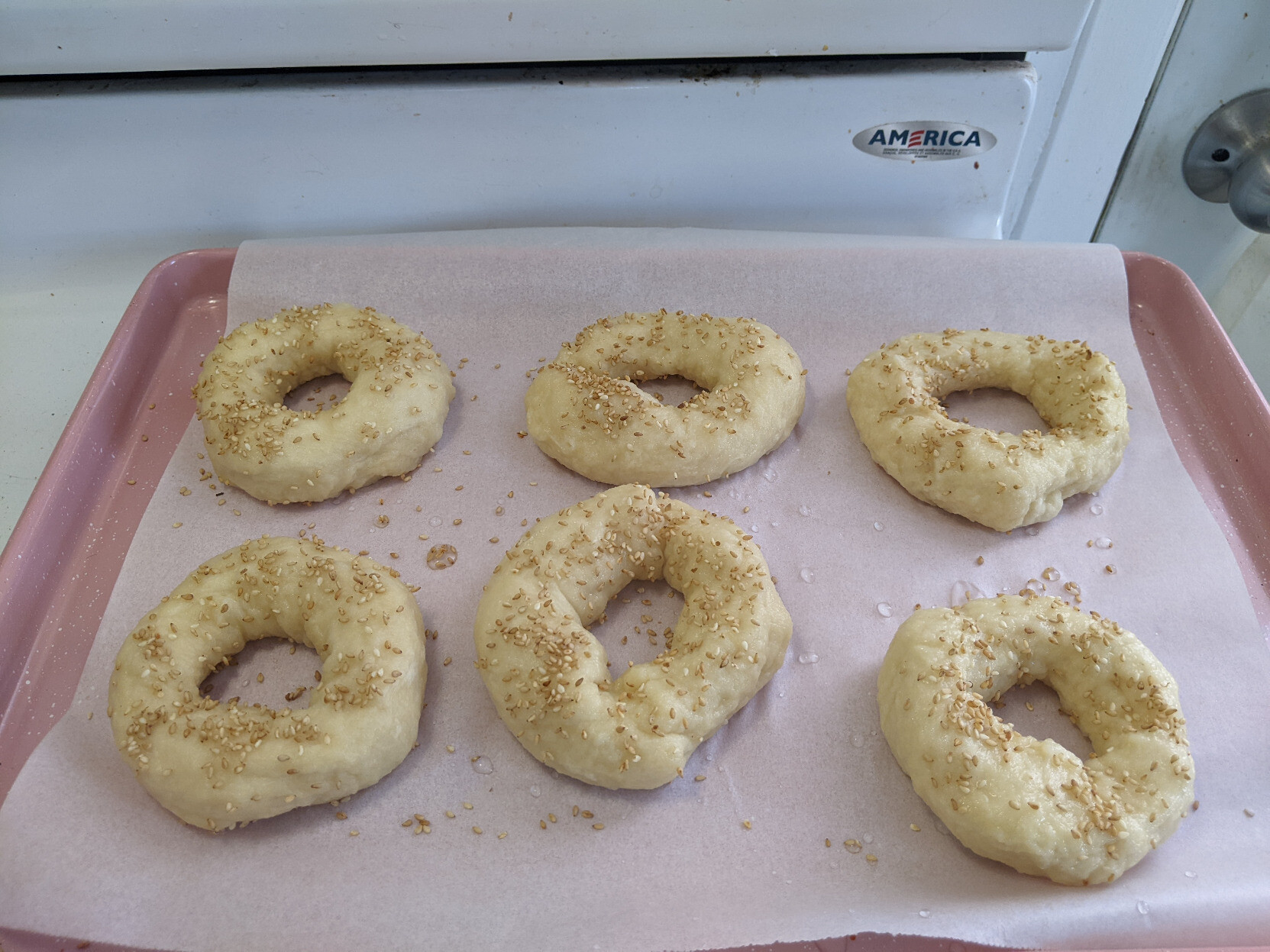 Unbaked homemade bagels on parchment paper on a pink baking wrap, sprinkled with light sesame seeds