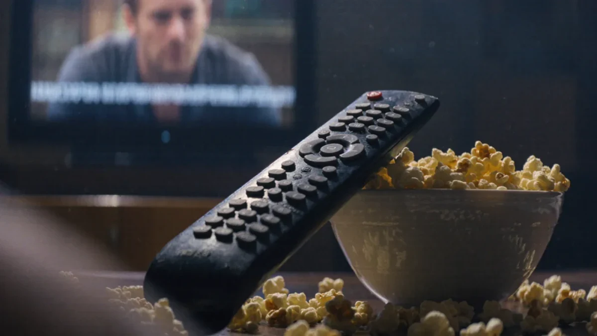Close-up of a remote, popcorn bowl, and television showing subtitles in a dark room