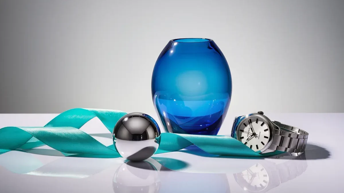 Still life of an electric blue glass vase, chrome sphere, silver watch, and turquoise silk ribbon on a pale gray surface